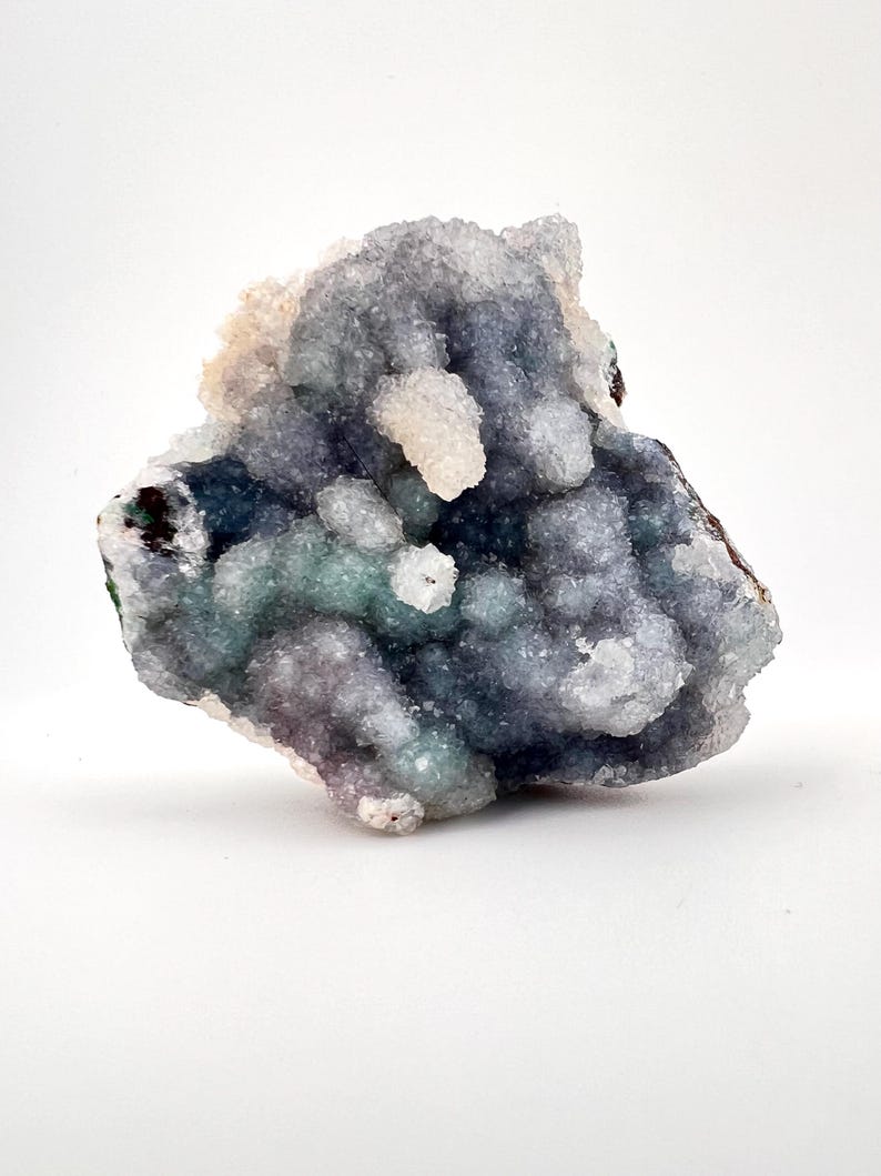 Puede incluir: Un grupo de cristales de cuarzo drusa en tonos de azul, verde y blanco. La formaci&oacute;n cristalina tiene una superficie rugosa y texturizada y se encuentra sobre un fondo blanco. El esp&eacute;cimen mineral muestra una forma natural y org&aacute;nica.