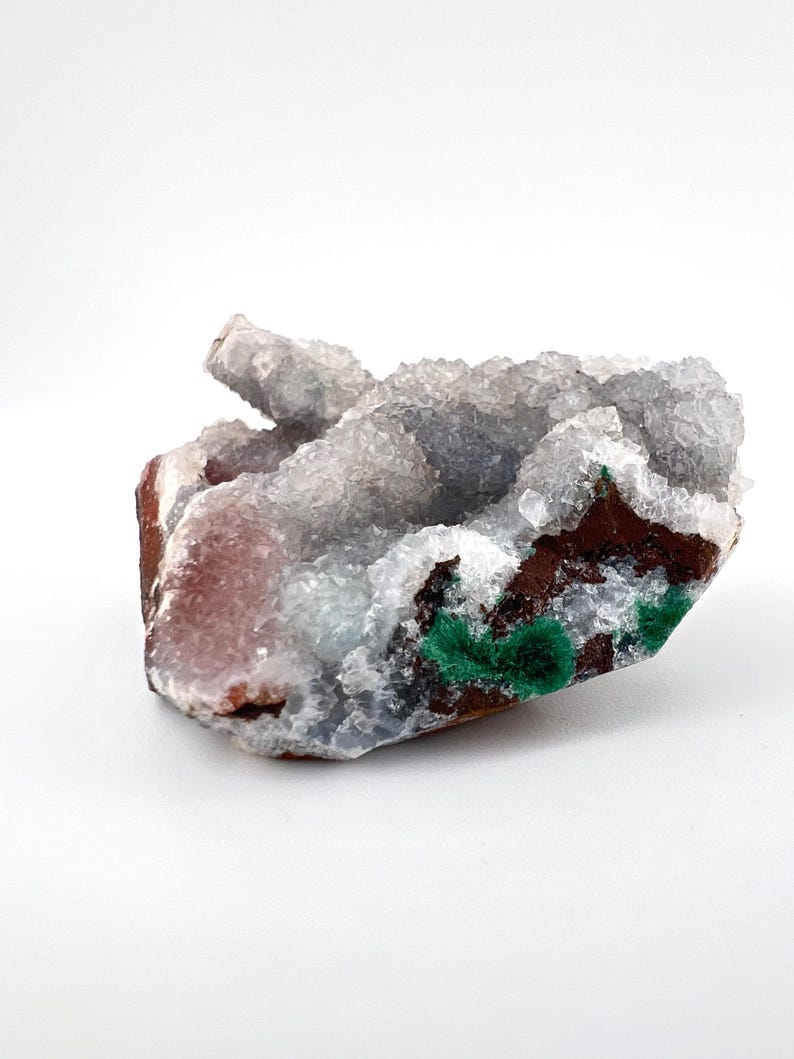 Puede incluir: Un c&uacute;mulo de cristales blancos, grises y rosados con un acento de malaquita verde. Los cristales est&aacute;n agrupados en una forma natural y rugosa.