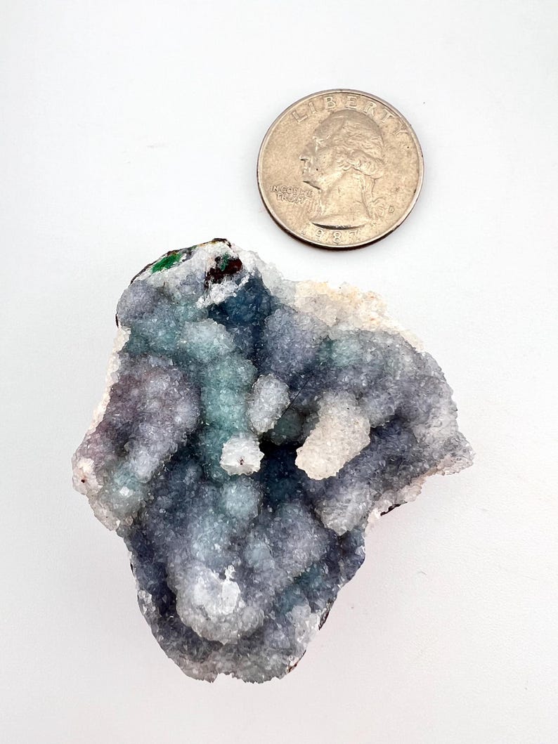 Puede incluir: Un grupo de formaciones cristalinas azules, verdes y blancas. Los cristales tienen una textura burbujeante y est&aacute;n junto a una moneda de veinticinco centavos para comparar el tama&ntilde;o. Los cristales est&aacute;n sobre un fondo blanco.