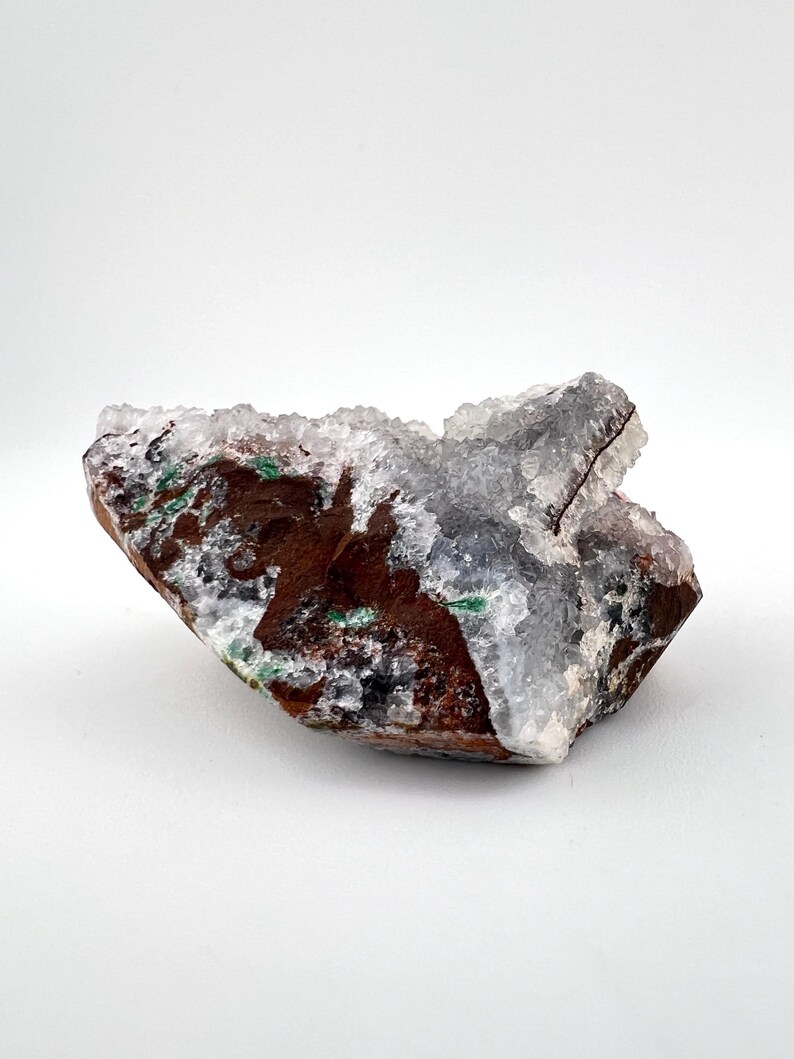 Puede incluir: Un c&uacute;mulo de cristales blancos transl&uacute;cidos con una matriz marr&oacute;n, verde y negra. Los cristales est&aacute;n agrupados en una forma irregular y rugosa.