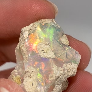 May include: A rough, white opal with iridescent flashes of green, orange, and red. The opal is embedded in a matrix of gray rock.