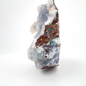 Puede incluir: Un esp&eacute;cimen de geoda natural con un exterior rugoso y texturizado. La geoda presenta una mezcla de colores, incluyendo tonos de marr&oacute;n, verde y azul-gris, con formaciones cristalinas. La geoda se muestra sobre un fondo blanco.