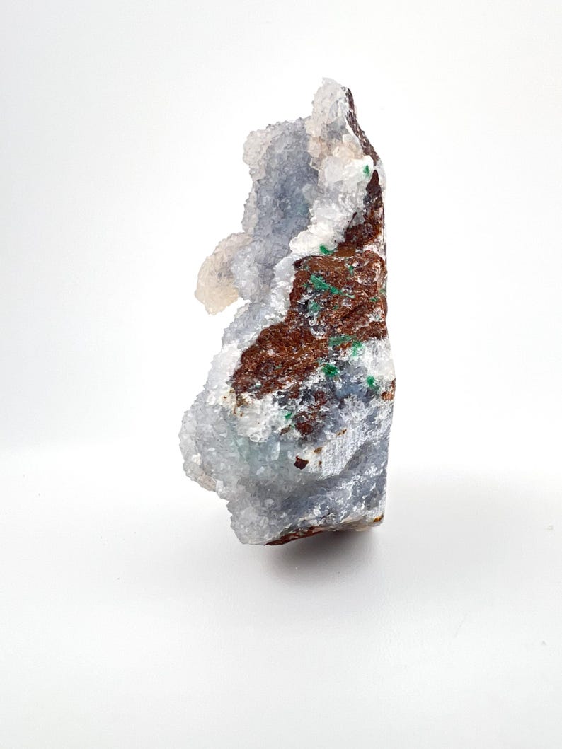 Puede incluir: Un esp&eacute;cimen de geoda natural con un exterior rugoso y texturizado. La geoda presenta una mezcla de colores, incluyendo tonos de marr&oacute;n, verde y azul-gris, con formaciones cristalinas. La geoda se muestra sobre un fondo blanco.