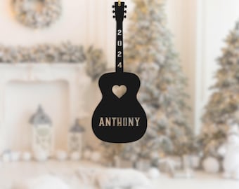 May include: Black guitar-shaped ornament with a heart cutout and the name "ANTHONY" in a serif font. The year "2024" is vertically displayed on the neck. A gold string is attached for hanging. The ornament is designed for holiday decor.