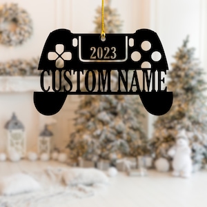 May include: Black Christmas ornament shaped like a video game controller. The ornament has the year "2023" and the words "CUSTOM NAME" cut out. A gold string is attached for hanging. The background is blurred with Christmas trees.