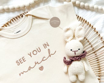 May include: A cream-colored baby onesie with the text "See you in March" embroidered in brown thread. A small heart is embroidered below the text. A crocheted white bunny with a brown scarf is sitting on the onesie. The onesie has a small brown label with the text "croissant label" on it.