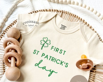 Könnte beinhalten: Ein cremefarbener Baby-Strampler mit einem grünen Kleeblatt und dem Text "FIRST ST PATRICK'S day", der auf der Vorderseite gestickt ist. Der Strampler liegt in einem geflochtenen Korb mit einer Holzrassel und einem Schnuller.