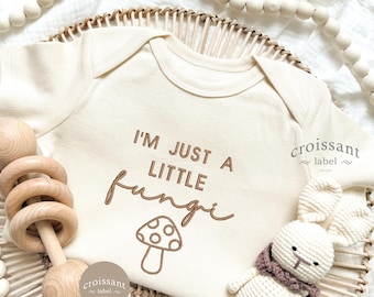 Peut inclure: Body de bébé crème avec le texte "I'M JUST A LITTLE fungi" et un dessin de champignon. Un hochet en bois et un lapin crocheté blanc avec une écharpe violette sont à côté du body. Le body est sur un panier tressé.