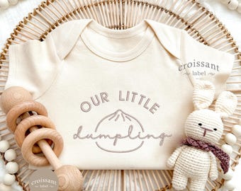 May include: Cream-colored baby onesie with the embroidered text "OUR LITTLE Dumpling" and a dumpling graphic. A wooden rattle and a crocheted bunny with a scarf are next to the onesie. The background includes a woven basket and wooden beads.