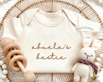 Może przedstawiać: Kremowe body dla dziecka z napisem "abuela's bestie" w brązowym skrypcie. Drewniana grzechotka i mały szydełkowy króliczek z fioletowym szalikiem leżą obok body. Tło obejmuje pleciony kosz i drewniane koraliki.