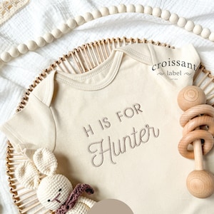 May include: A cream-colored baby onesie with the text "H is for Hunter" embroidered in brown thread. The onesie is lying on a woven wicker tray with a wooden rattle and a crocheted bunny toy.