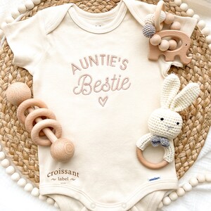 May include: A white baby onesie with the text "Auntie's Bestie" embroidered in pink. The onesie is surrounded by wooden toys, including a rattle, a bunny, and an elephant.