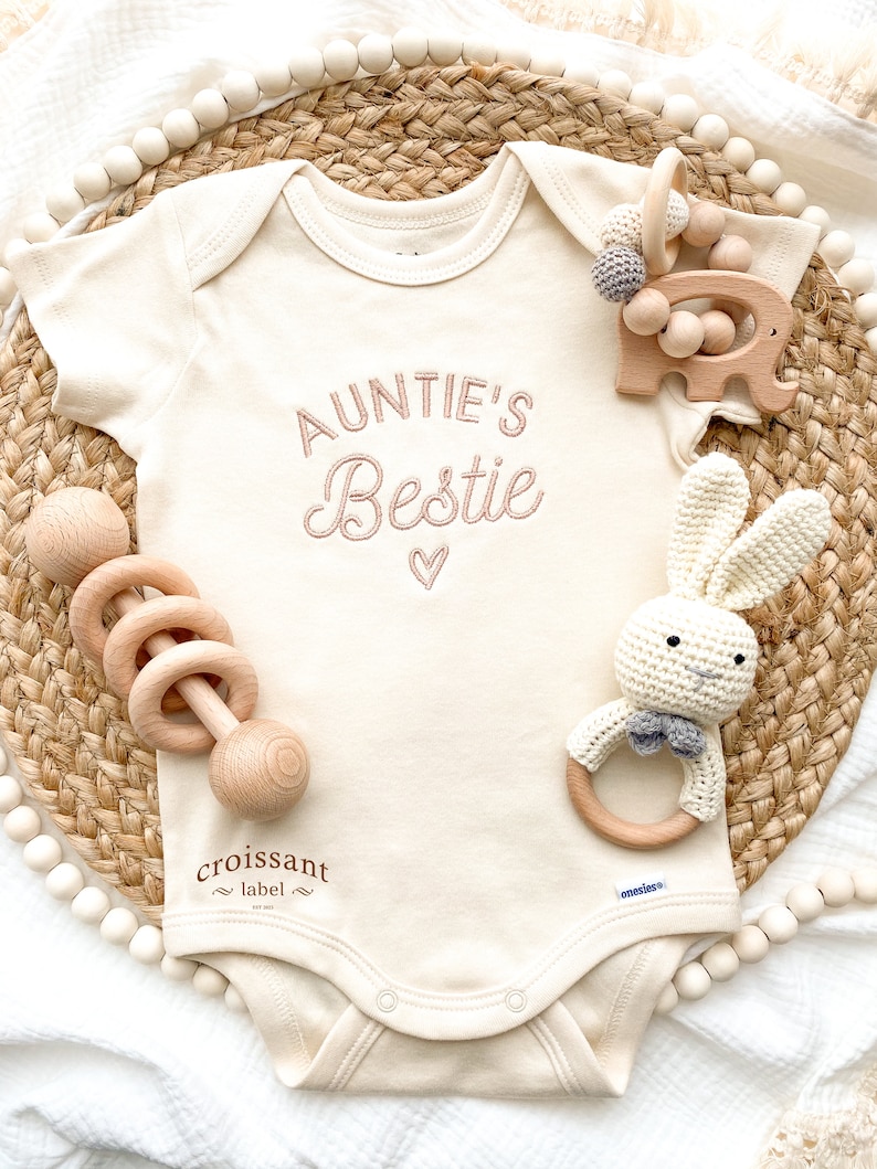May include: A white baby onesie with the text "Auntie's Bestie" embroidered in pink. The onesie is surrounded by wooden toys, including a rattle, a bunny, and an elephant.