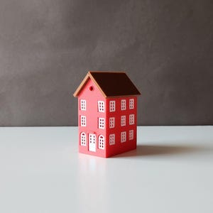 Puede incluir: Una pequeña casa de juguete roja con un techo marrón y ventanas blancas. La casa tiene una puerta y está sentada sobre una superficie blanca.