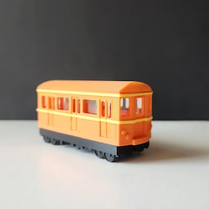Könnte beinhalten: Orangener Spielzeug-Zugwagen mit schwarzen Rädern und gelben Akzenten. Der Wagen hat Fenster und eine Tür.