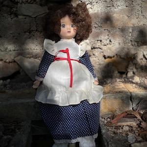May include: A vintage doll with brown curly hair and blue eyes. The doll wears a white apron with a red ribbon over a navy blue dress with white polka dots. The doll is seated on a stone surface.