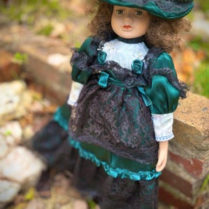 May include: A porcelain doll with brown curly hair, wearing a green satin dress with black lace overlay and a matching hat. The doll has a painted face with green eyes and red lips. The dress features white lace trim and teal ribbon accents.