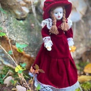 May include: A vintage doll dressed in a deep red velvet dress and matching hat. The doll has long brown braids and a white lace collar and cuffs. The doll is standing in a natural outdoor setting with leaves and greenery.