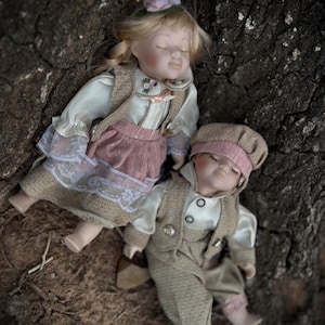 May include: Two porcelain dolls, a girl and a boy, resting on a textured, dark surface. The girl doll wears a pink skirt, a white blouse, and a green hat with a flower. The boy doll is dressed in a tweed suit and a matching cap.