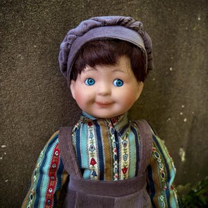 May include: A vintage porcelain doll with blue eyes and brown hair, wearing a striped shirt, corduroy overalls, and a matching cap. The doll has a serene expression and is set against a textured background.