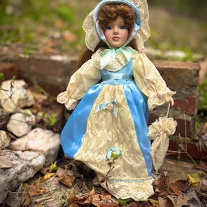 May include: A porcelain doll dressed in a blue and cream-colored dress with lace accents. The doll has brown hair, blue eyes, and a bonnet with lace trim. She holds a small parasol and a flower.