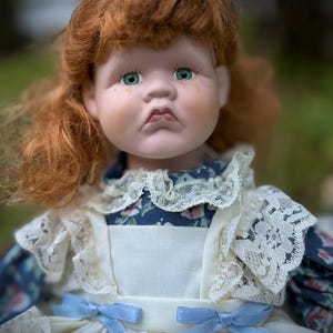 May include: A porcelain doll with red hair and green eyes. The doll wears a blue floral dress with a white apron and lace trim. Light blue ribbons adorn the dress. The doll has a sad expression with tear streaks.