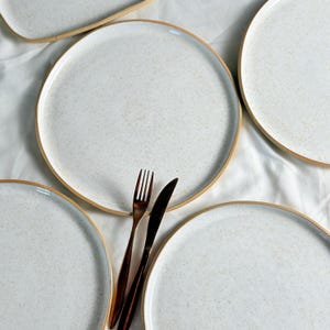 May include: Four white ceramic dinner plates with a speckled glaze and a brown rim. A gold fork and knife are placed on top of one of the plates.