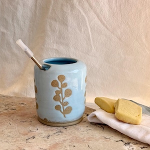 May include: A light blue ceramic cup with a speckled tan botanical leaf pattern and a notched rim. A silver metal straw with a white silicone tip is inserted into the notch. The cup has a smooth, glossy finish and a natural, unglazed tan base.