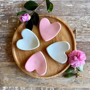 May include: Four heart-shaped ceramic dishes, two in pale pink and two in speckled light blue, arranged on a round wooden tray. Pink and green flowers and foliage are placed around the tray.