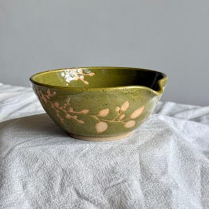 May include: A round ceramic mixing bowl with a glossy olive green glaze and a speckled finish. The exterior features a light pink botanical pattern of small leaves and branches. The bowl includes a small pouring spout on the right side rim.