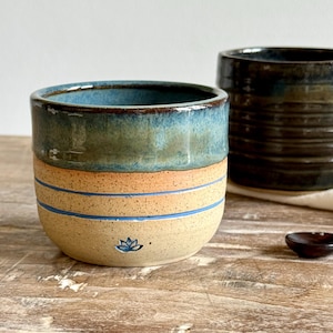 May include: A ceramic cup with a speckled beige lower half featuring two horizontal blue stripes and a small blue lotus flower icon. The upper half has a smooth, glossy blue-green glaze. A dark brown, ribbed ceramic cup sits in the background.