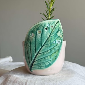Peut inclure: Un vase en céramique en forme de feuille, vert et blanc, avec une base rose clair. Le vase présente un motif de nervures de feuilles détaillé et plusieurs petits trous. Une brindille de plante verte est placée dans le vase.
