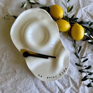 May include: A white ceramic serving dish with a distinctive, undulating shape. It features two small wooden spoons and a yellow spatula. The words "La Dolce Vita" are written in black script. Three yellow lemons and olive branches are arranged nearby.