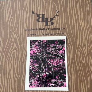 May include: A pack of 12 pink and black camouflage arrow wraps, 4 inches in length. The wraps are printed with a pink and black camouflage pattern. The packaging features the text "Bucks & Bulls Trading Co." and "12 pack 4 inch Arrow Wraps".