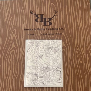 May include: A brown paper background with a black and white logo of a buck and bull head with the text "Bucks & Bulls Trading Co." and "12 pack 4 inch Arrow Wraps". A white rectangle with a black and white topographic map design is in the center of the image.