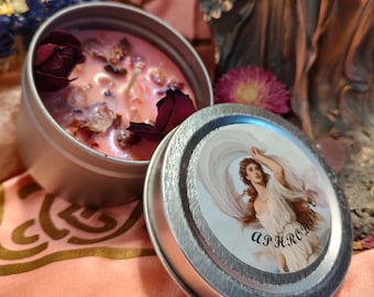 Puede incluir: Una vela rosa con pétalos de rosa secos y cristales en una lata de plata. La tapa de la lata presenta una ilustración vintage de una mujer con la palabra "APHRODITE" escrita debajo de ella.