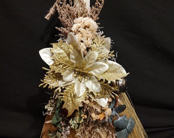 May include: A decorative broom with a handle wrapped in cream-colored fabric. The broom is adorned with gold and white flowers, dried foliage, and a small silver bell. The broom's base is made of straw and feathers.