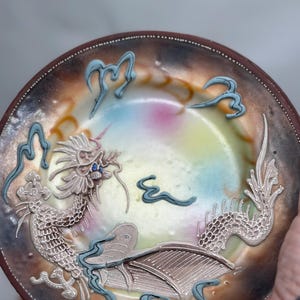 Può includere: Un piatto decorativo con un design dettagliato di drago. Il piatto presenta un drago in rilievo argentato e blu, su uno sfondo colorato con un bordo marrone. Il drago ha squame intricate e un occhio di pietra preziosa blu.
