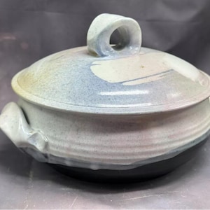 Puede incluir: Una fuente de cerámica con tapa y asa de bucle. La tapa y el cuerpo son blancos con detalles en azul, amarillo y negro. La base es negra. El plato tiene dos asas laterales.