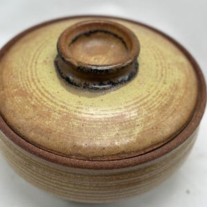 May include: A small, round, ceramic bowl with a lid. The bowl is a light brown color with a darker brown rim. The lid has a small, round knob on top.