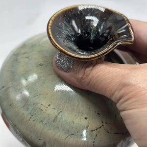 Puede incluir: Un jarrón de cerámica hecho a mano con una forma orgánica única. El jarrón presenta un cuerpo verde moteado y un borde acampanado marrón oscuro. El borde tiene un acabado brillante y vidriado, lo que agrega un toque de elegancia. El jarrón es sostenido por una persona.
