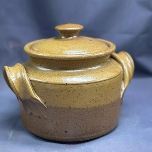 Puede incluir: Un tarro de cerámica marrón con tapa y dos asas. El tarro tiene un esmalte moteado y una banda marrón más oscura alrededor del centro.