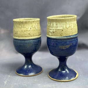 May include: Two handmade ceramic goblets with a blue and beige glaze. The goblets have a wide bowl and a tall stem.
