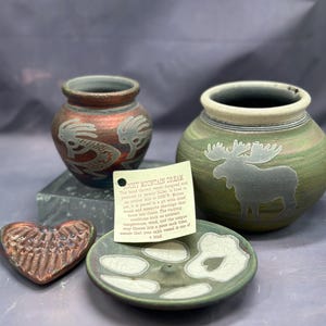 May include: A collection of handmade pottery pieces. Includes a small red-brown vase with a gray design, a green vase with a moose silhouette, a heart-shaped piece, and a small dish. The pieces have a rustic, earthy aesthetic.