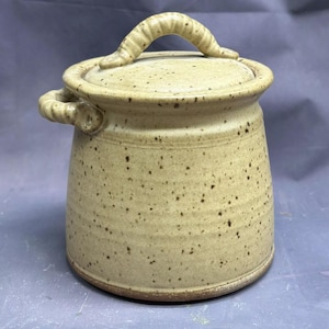 Pode incluir: Um pote de cerâmica bege com manchas, com tampa e uma alça torcida. O pote tem um esmalte bege com manchas.