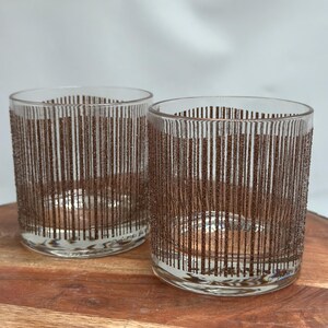 May include: Two clear drinking glasses with a brown vertical stripe design. The glasses are cylindrical and sit on a wooden surface. The glasses are empty and appear to be made of glass.