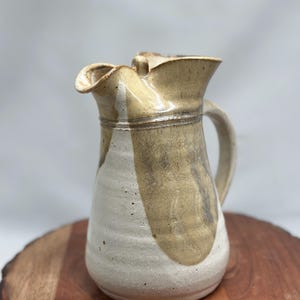 Może przedstawiać: Ręcznie robiony ceramiczny dzbanek z kremowym i jasnobrązowym szkliwem. Dzbanek ma unikalny, falisty brzeg i zakrzywiony uchwyt. Projekt przedstawia jasnobrązowy wzór wirowy na kremowej podstawie. Dzbanek stoi na drewnianej powierzchni.