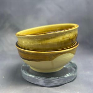 May include: Two ceramic bowls stacked on top of each other. The top bowl is a pale yellow with a brown rim. The bottom bowl is a pale yellow with brown streaks.