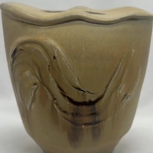 Könnte beinhalten: Eine hellbraune Keramikvase mit einem wellenförmigen Design. Die Vase hat eine breite Öffnung und einen abgerundeten Boden.