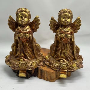 Puede incluir: Dos figuras de querubines dorados sosteniendo liras, cada una sentada sobre una base dorada con detalles intrincados. Las bases están unidas a un tronco de madera.